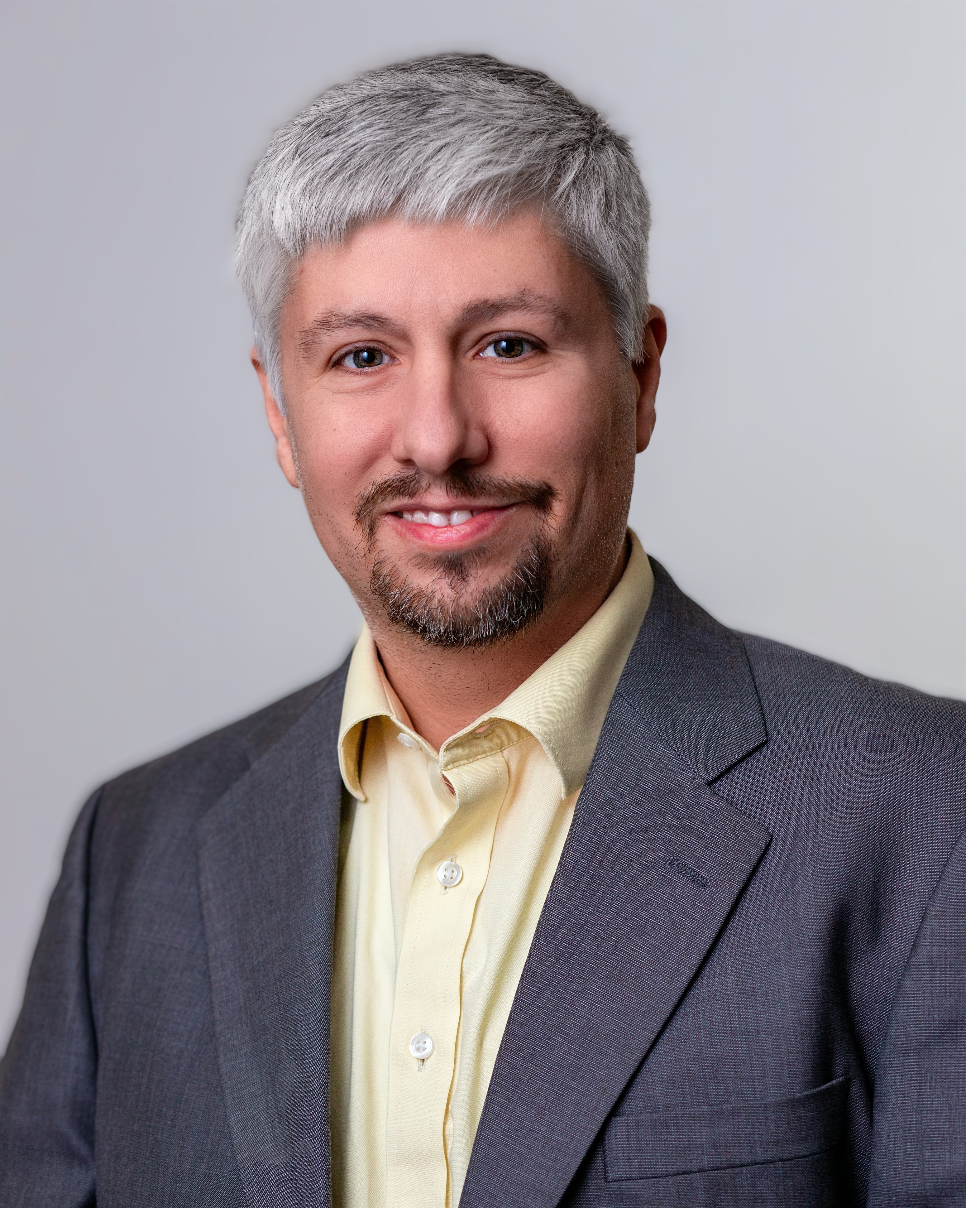 a smiling man with grey hair wearing a yellow shirt and a dark grey suit jacket