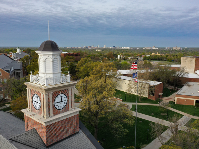 Overhead photo of campus with a clocktower and flag pole with the United States and Kansas flags in the foreground and the Wichita downtown skyline in the background