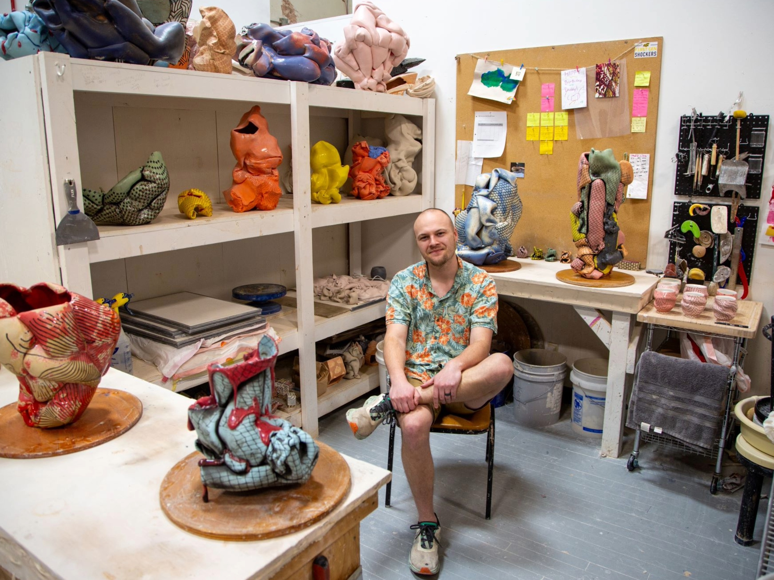 MFA student Scott Jones sits in a chair in his Henrion Hall studio surrounded by art supplies and multicolored ceramic sculptures.