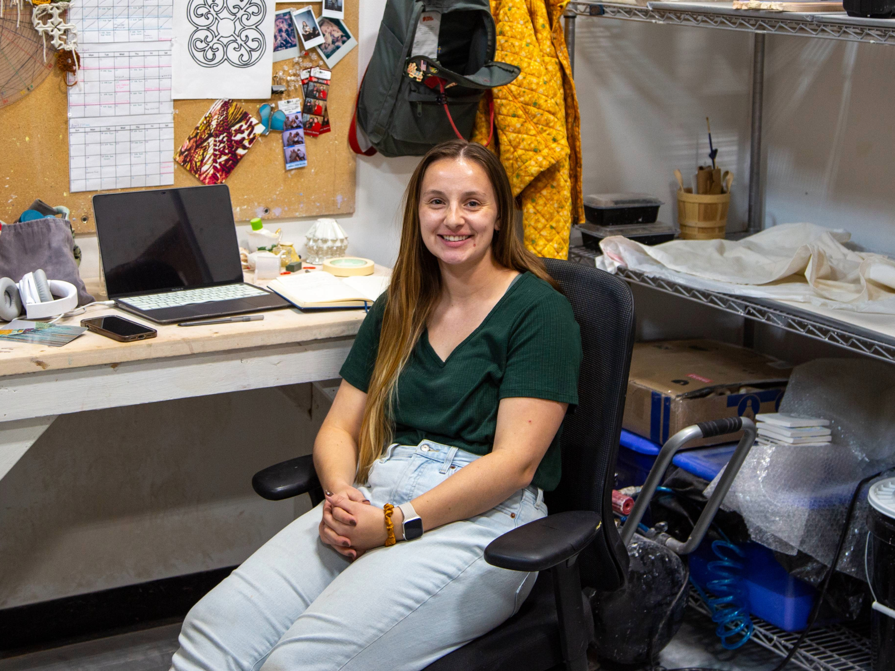 MFA student Mollykate Geddis sits in a desk chair in her Henrion Hall studio surrounded by art supplies.
