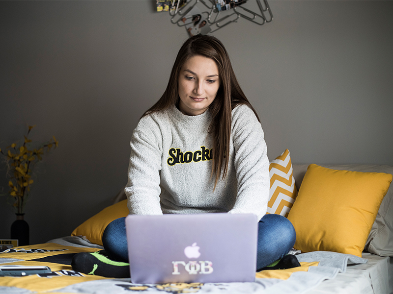 A student sitting in their room and looking at a laptop.