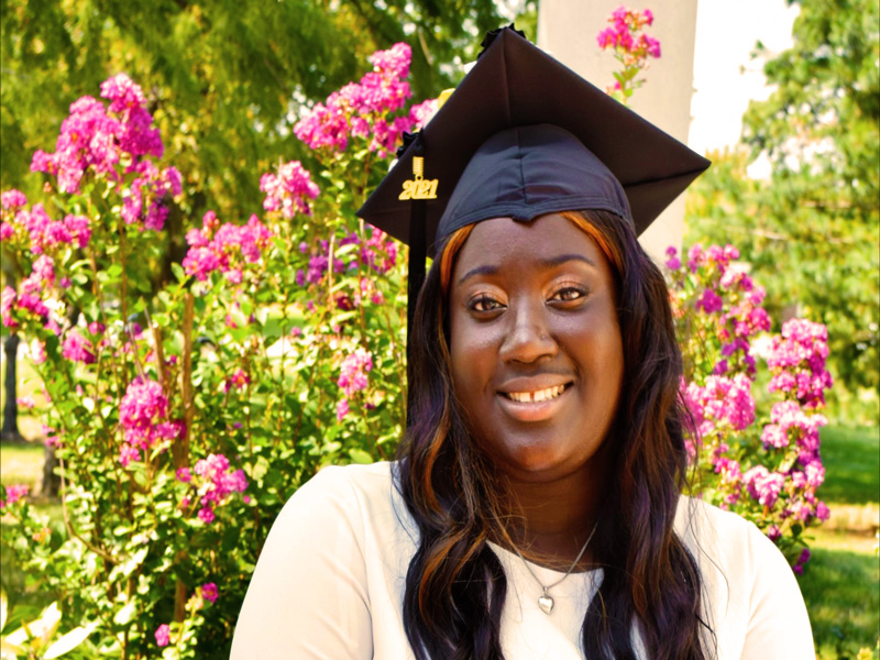 Aunjnae sits in front of purple-pink flowery bush. She has her graduation cap on and is wearing a white blouse and a silver heart locket. 