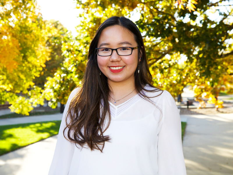 Joyce Chiam standing in front of the steps of Ablah library outlooking campus with lush green and yellow trees in the background. She has a white blouse, long dark hair, black frame glasses, and coral red lipstick on. 