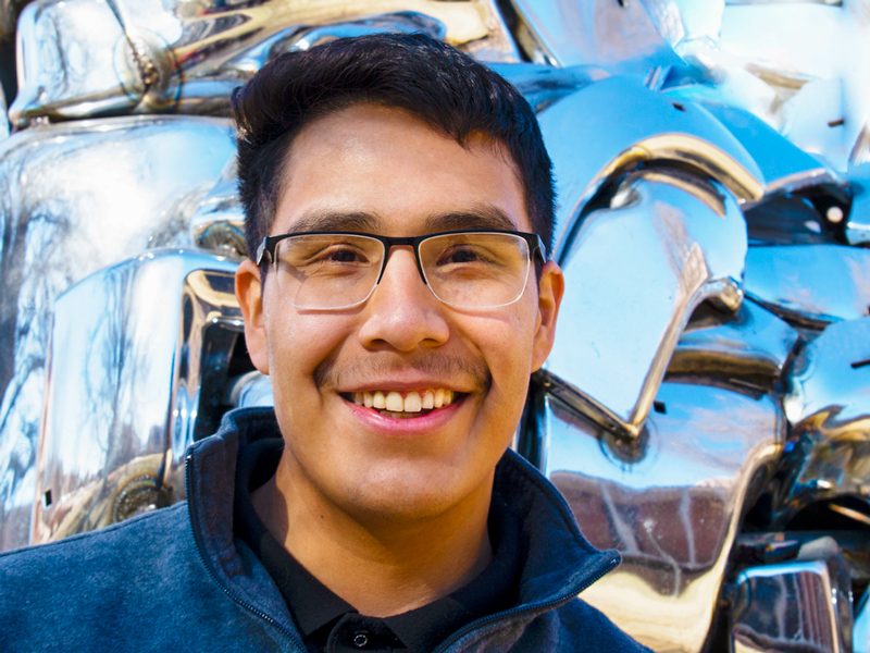 Juan Aguilar is standing outside behind a metal sculpture. 