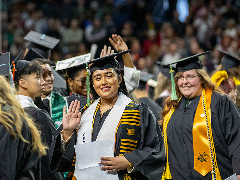 A Wichita State University graduate waves at the camera, surrounded by other graduates. 