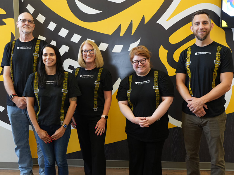 Members of the physician associate program pose in front of a wall with Wu on it. Members of the program are all wearing shirts that read "Suspenders4Hope #WeSupportU," with stylized yellow suspenders that are made up of different words printed on the shirts.