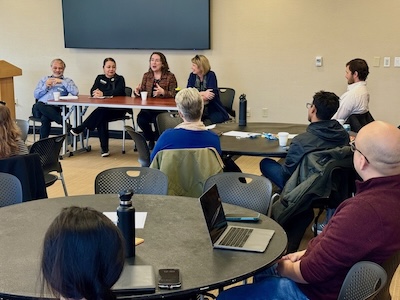 Four faculty sit at a table at the front of the room giving a panel at New Faculty On-Ramp. New faculty sit around tables looking on.
