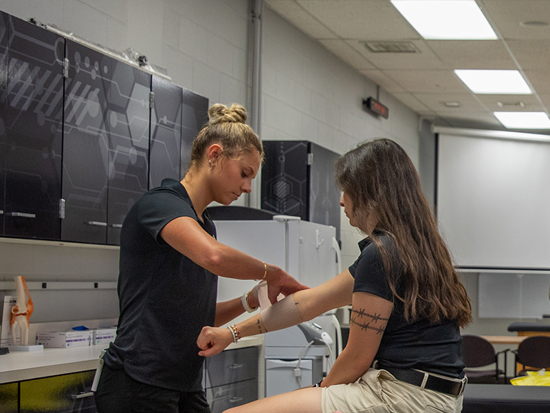 One student wraps a bandage on another's arm.