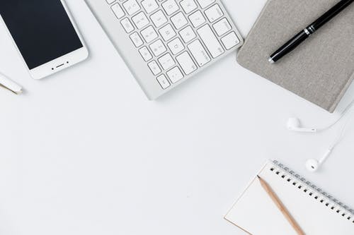 Photo of a mobile phone, a computer keyboard, a journal, and a pocket notebook on a white background or table. 