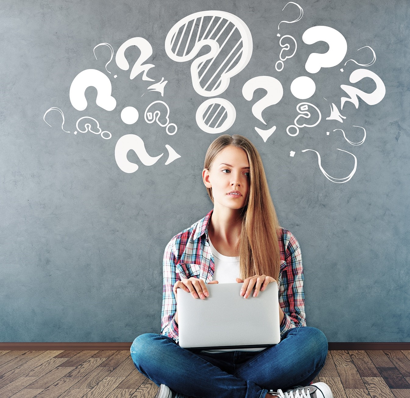 A young woman with a computer sitting in front of a blackboard. The blackboard has many questions marks drawn in a way that make it seem as if they're emerging from her head. 