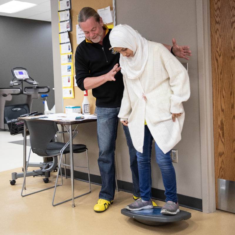 Trainer assisting participant in balance test on a wobble board during a physical assessment.
