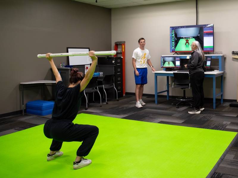 A woman performs a squat with a bar overhead on a green mat, while two observers monitor her form on screens displaying DEXA motion analysis.