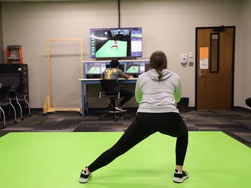 Woman performing a lunge on a green mat using the DARI motion-capture system, monitored by a man at a computer workstation.