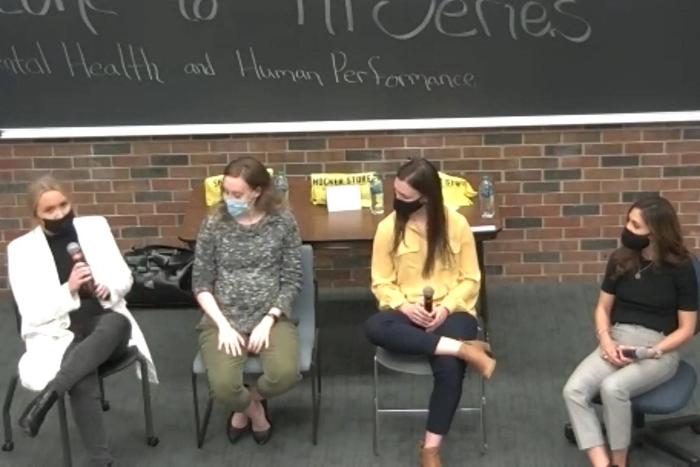 Four women seated on a panel in front of a chalkboard reading 'Mental Health and Human Performance,' with three of them holding microphones.