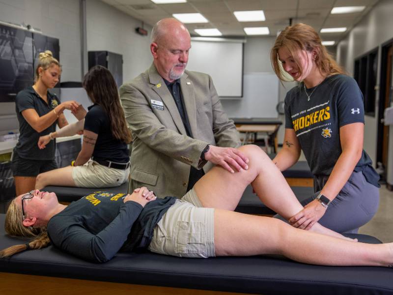 A student reclines on a physical therapy table in the Human Performance lab while faculty and fellow students examine her knee.