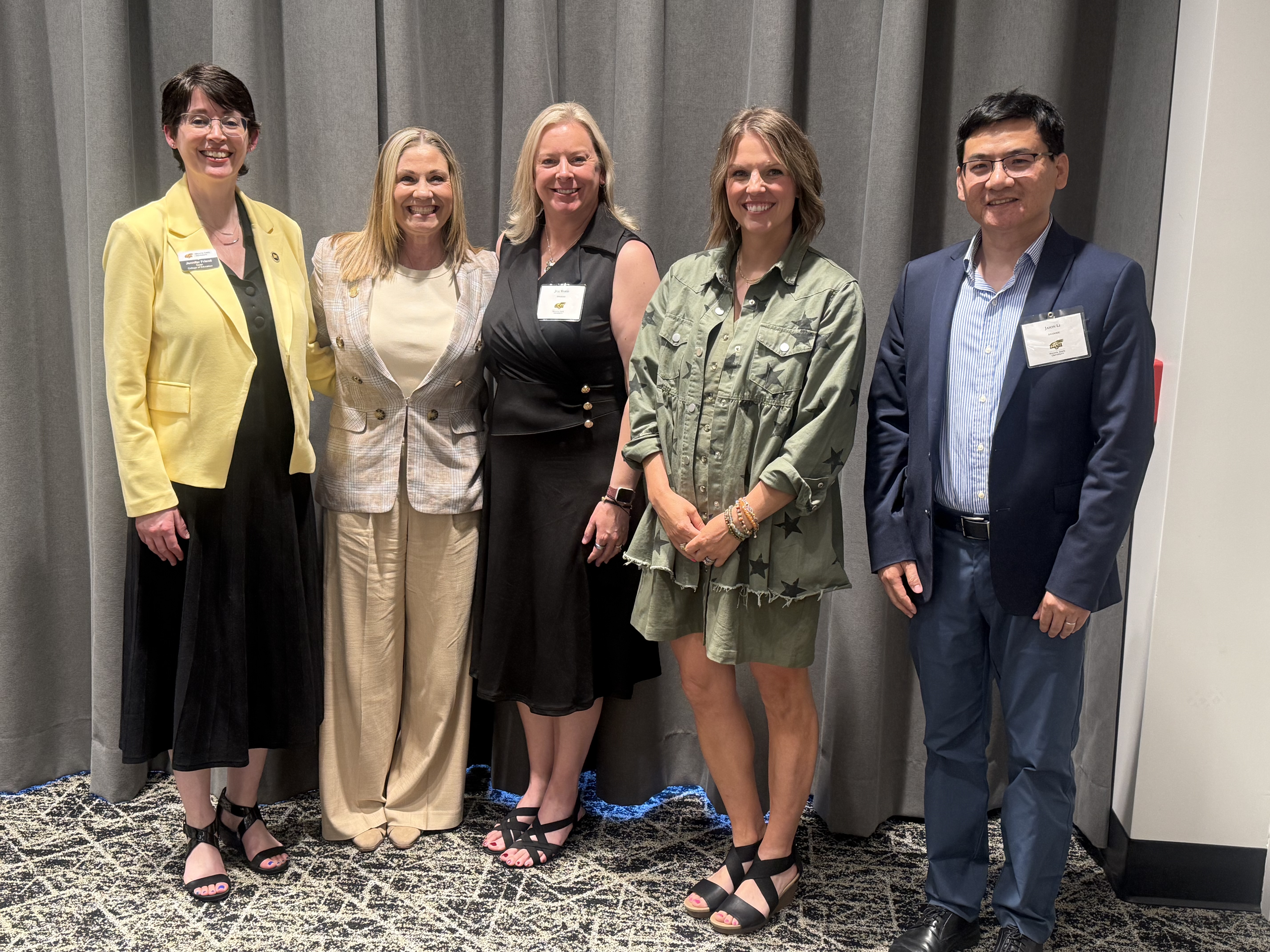 Five individuals stand together smiling for a photo in front of gray curtains during a College of Education tenure and promotion recognition event. The group includes faculty and staff members dressed in professional attire, including suits, dresses and business casual clothing, all wearing name badges.