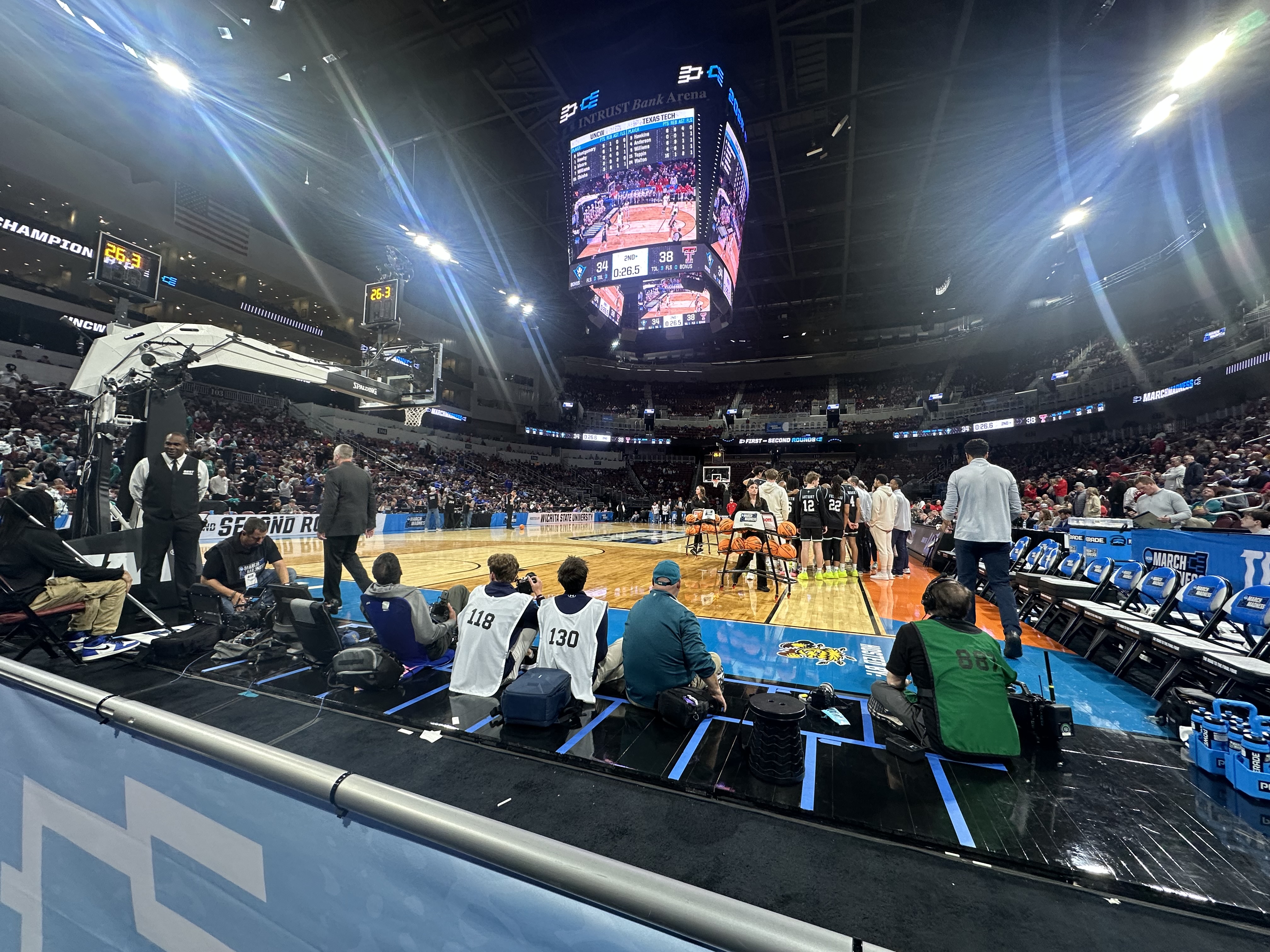 A view from courtside at the NCAA men’s basketball tournament in Wichita. The arena is filled with fans, and players gather near the bench during a timeout. Media and camera crews are visible along the baseline.