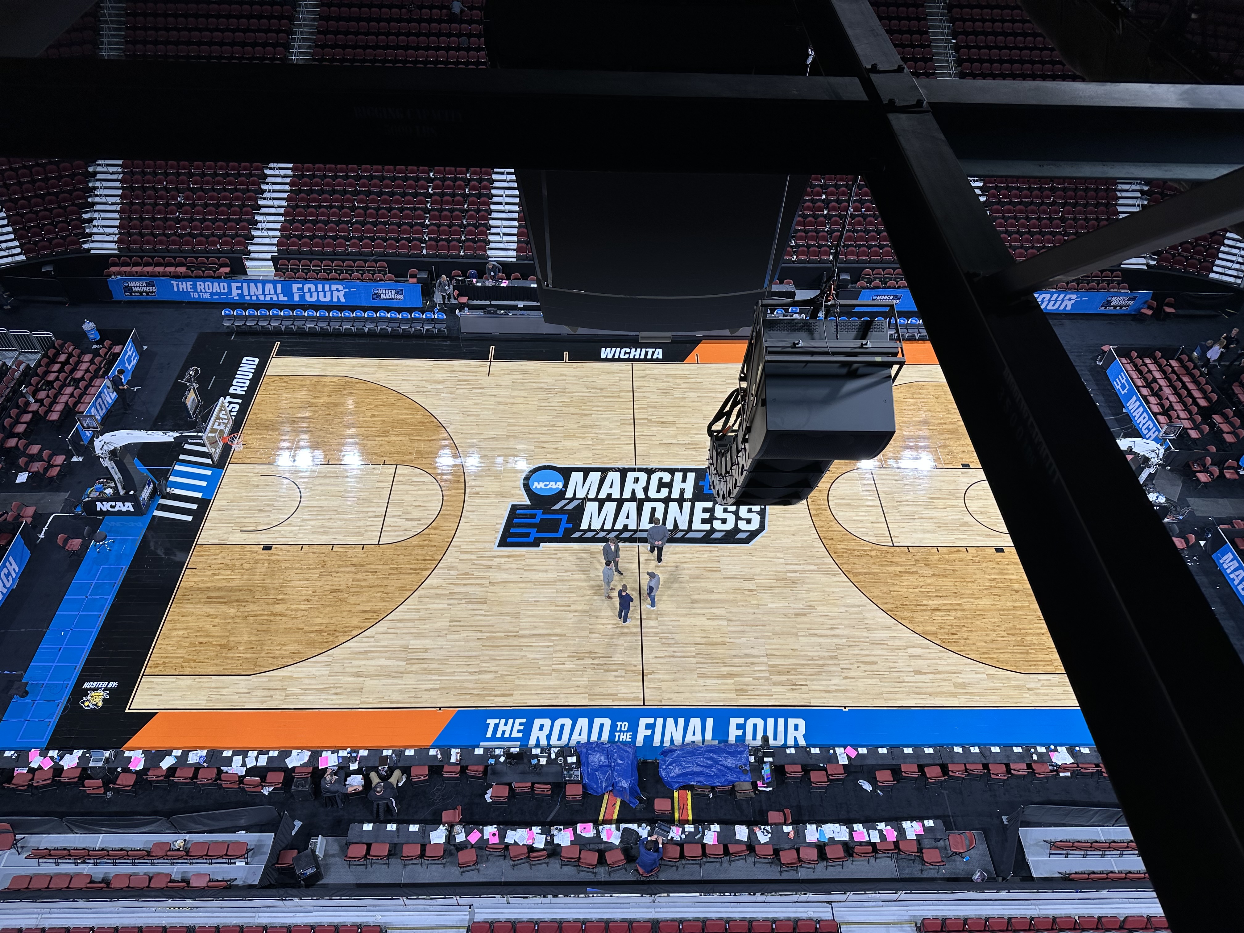 A high-angle photo of an NCAA basketball court taken from the catwalk area above the arena. The March Madness logo is centered on the court with setup crews visible below.