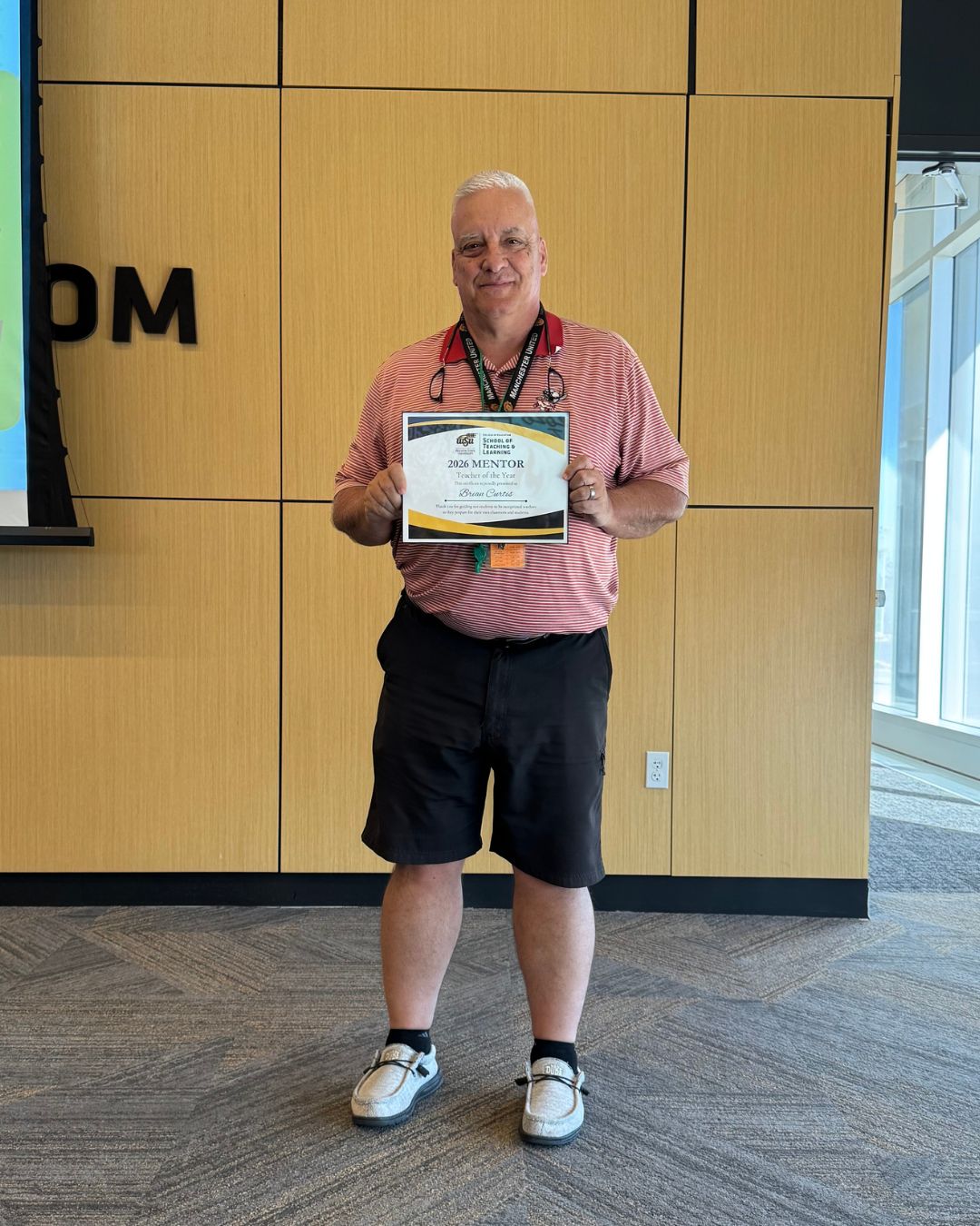 A man stands indoors holding a certificate that reads “2026 Mentor Teacher of the Year” from Wichita State University’s School of Teaching and Learning. He is wearing a red striped polo shirt, black shorts and gray shoes, standing in front of a wood-paneled wall.
