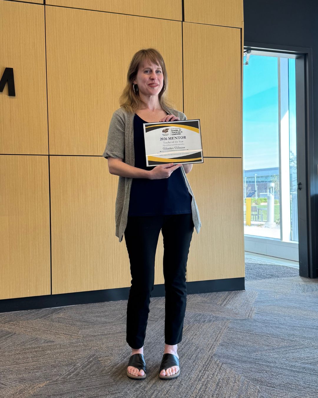 A woman stands indoors holding a “2026 Mentor Teacher of the Year” certificate from Wichita State University’s School of Teaching and Learning. She is wearing a black top, gray cardigan and black pants, standing near a window with natural light.
