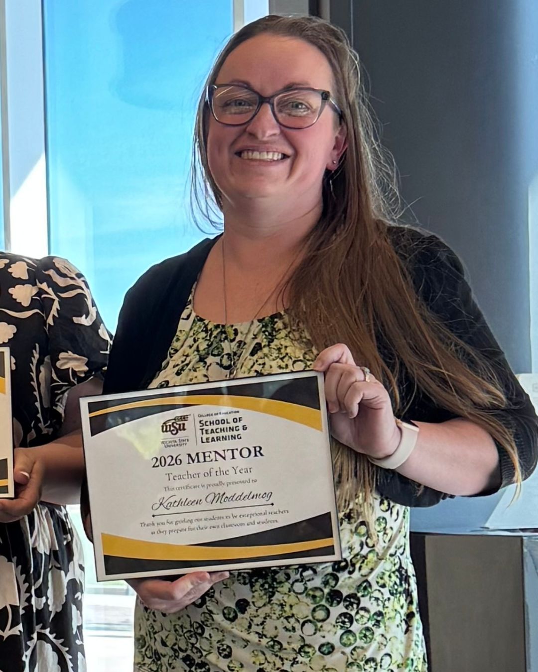 A woman smiles while holding a “2026 Mentor Teacher of the Year” certificate from Wichita State University’s School of Teaching and Learning. The photo is framed closer, showing her from the chest up with a window in the background.