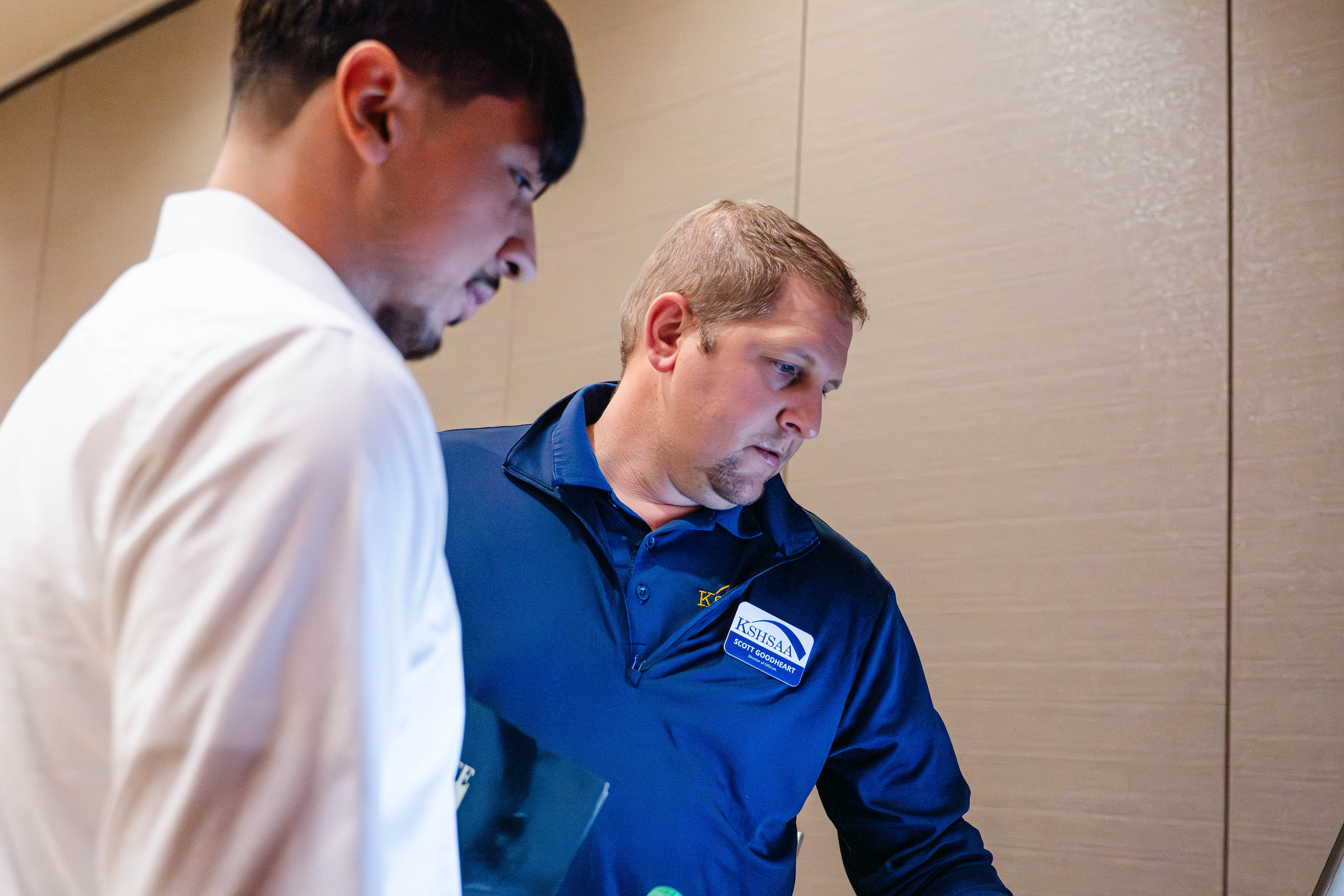 Two men review information at a booth during the Kansas Sport Summit at Wichita State University, looking down at materials together in a conference room setting.