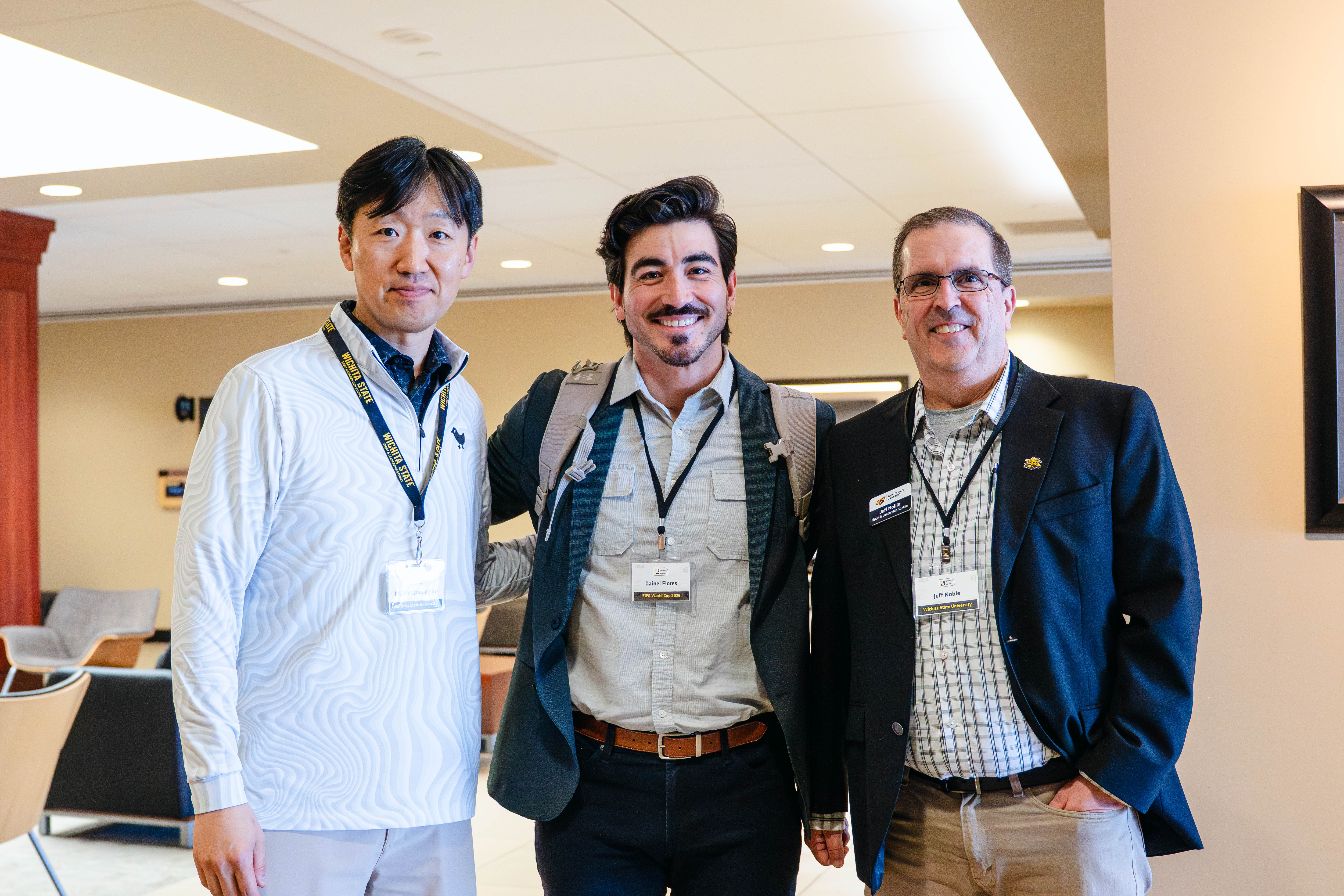 Two men review information at a booth during the Kansas Sport Summit at Wichita State University, looking down at materials together in a conference room setting.  Three men pose together at the Kansas Sport Summit at Wichita State University, smiling and wearing name badges in a lobby area.