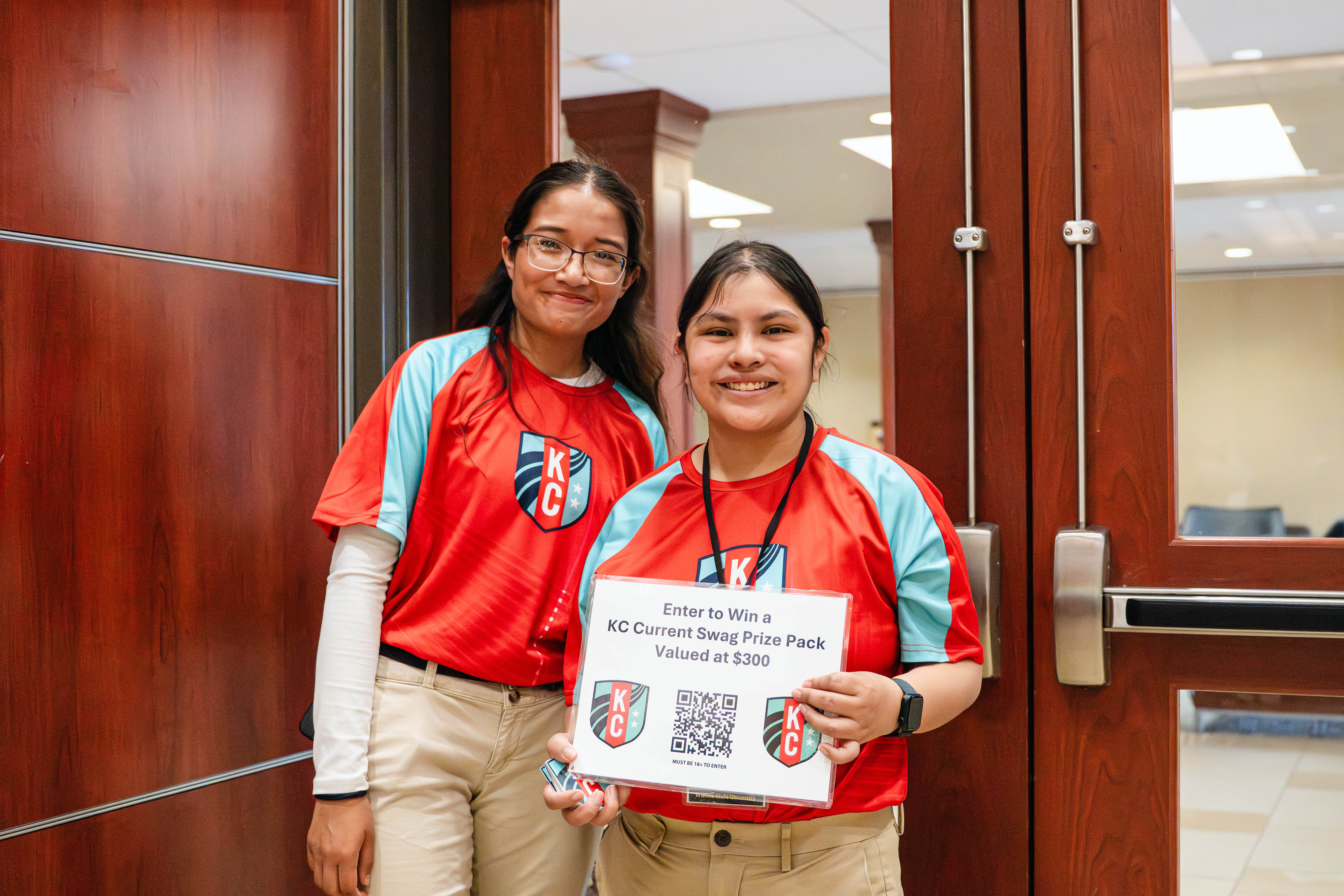 Two individuals stand at an event entrance during the Kansas Sport Summit at Wichita State University, holding a sign promoting a Kansas City Current giveaway.