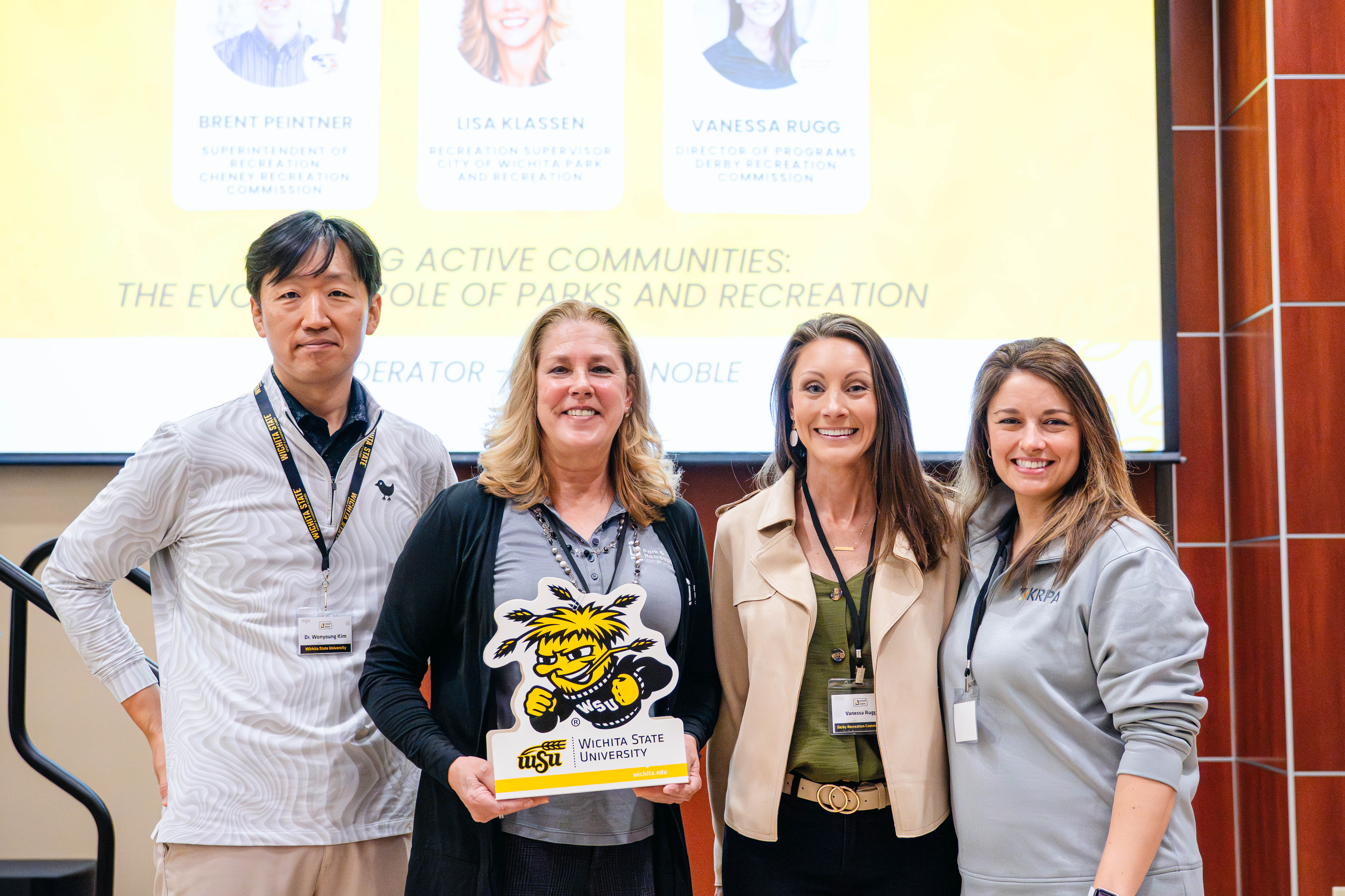 Four presenters pose together in front of a presentation screen during a panel session at the Kansas Sport Summit at Wichita State University.