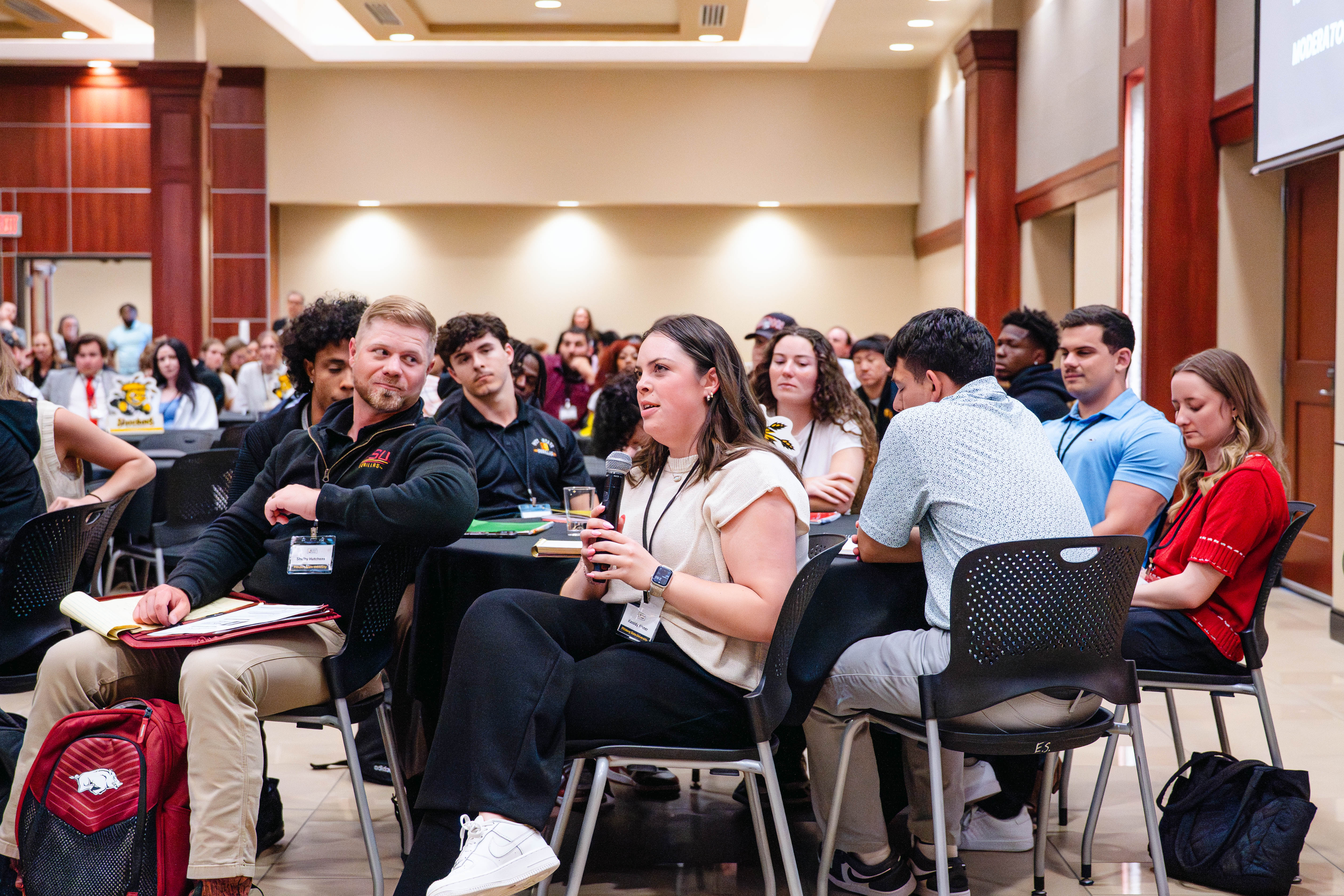 An attendee holds a microphone and asks a question while seated at a table during a session at the Kansas Sport Summit at Wichita State University.