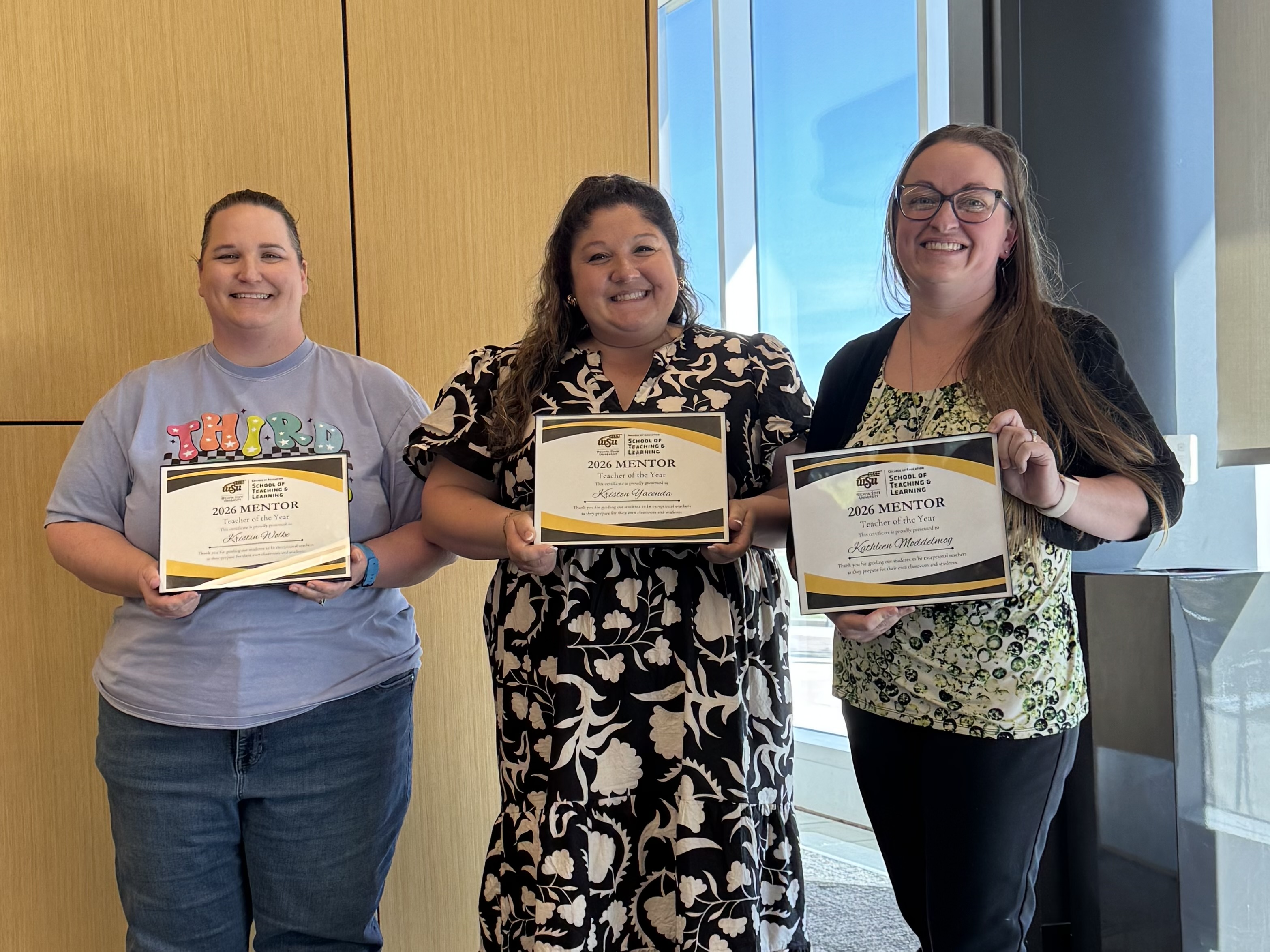 Three women stand indoors holding “2026 Mentor Teacher of the Year” certificates from Wichita State University’s School of Teaching and Learning. They are smiling and posed side by side in front of a wood-paneled wall with a window and natural light to the right.