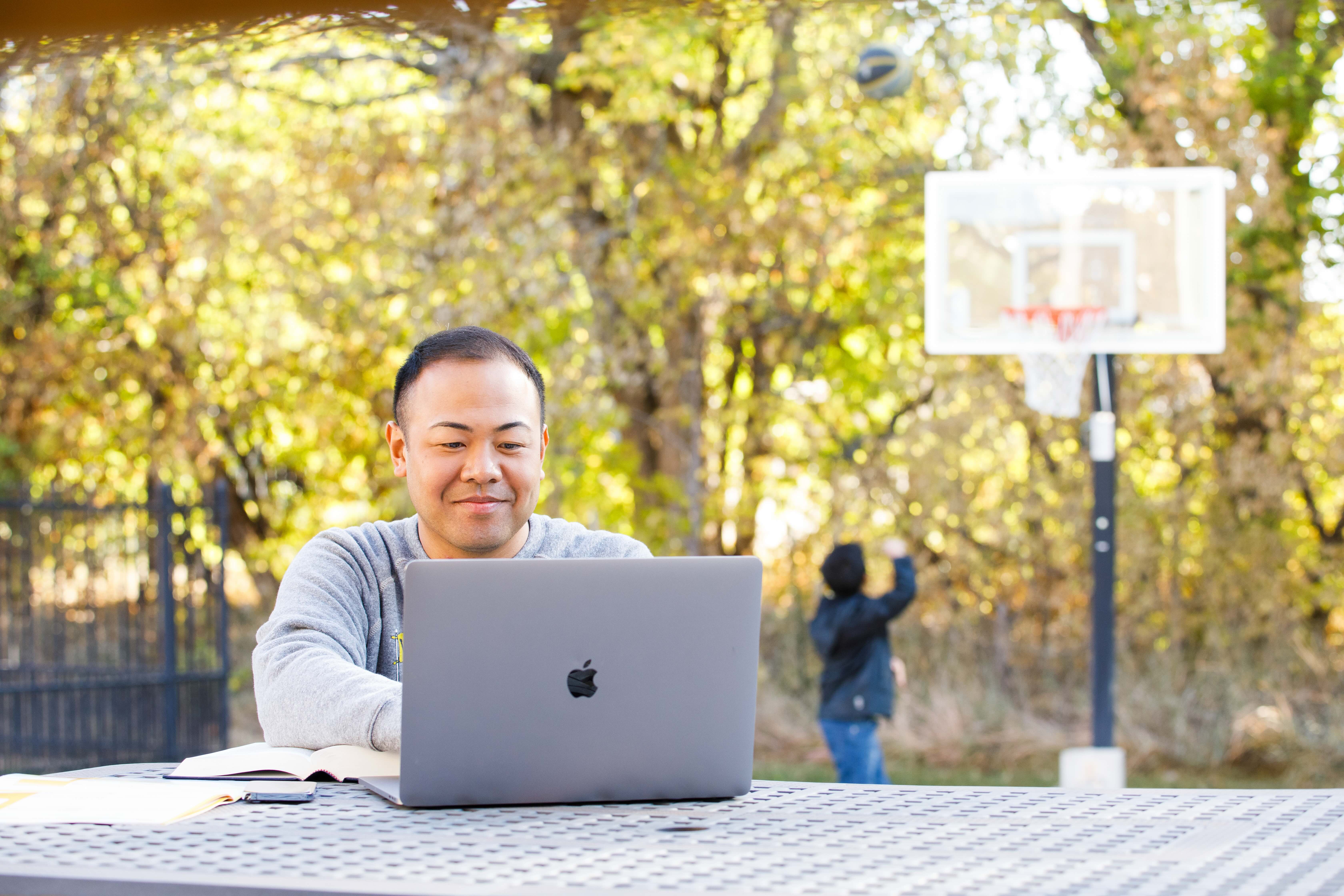 Student on laptop at a table outdoors with trees in the background, child in background with basketball goal, online student