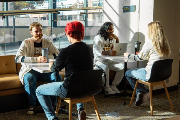students on campus at wichita state university sitting at tables facing each other, two per table, with laptops and coffee mugs, talking and studying with windows in the background