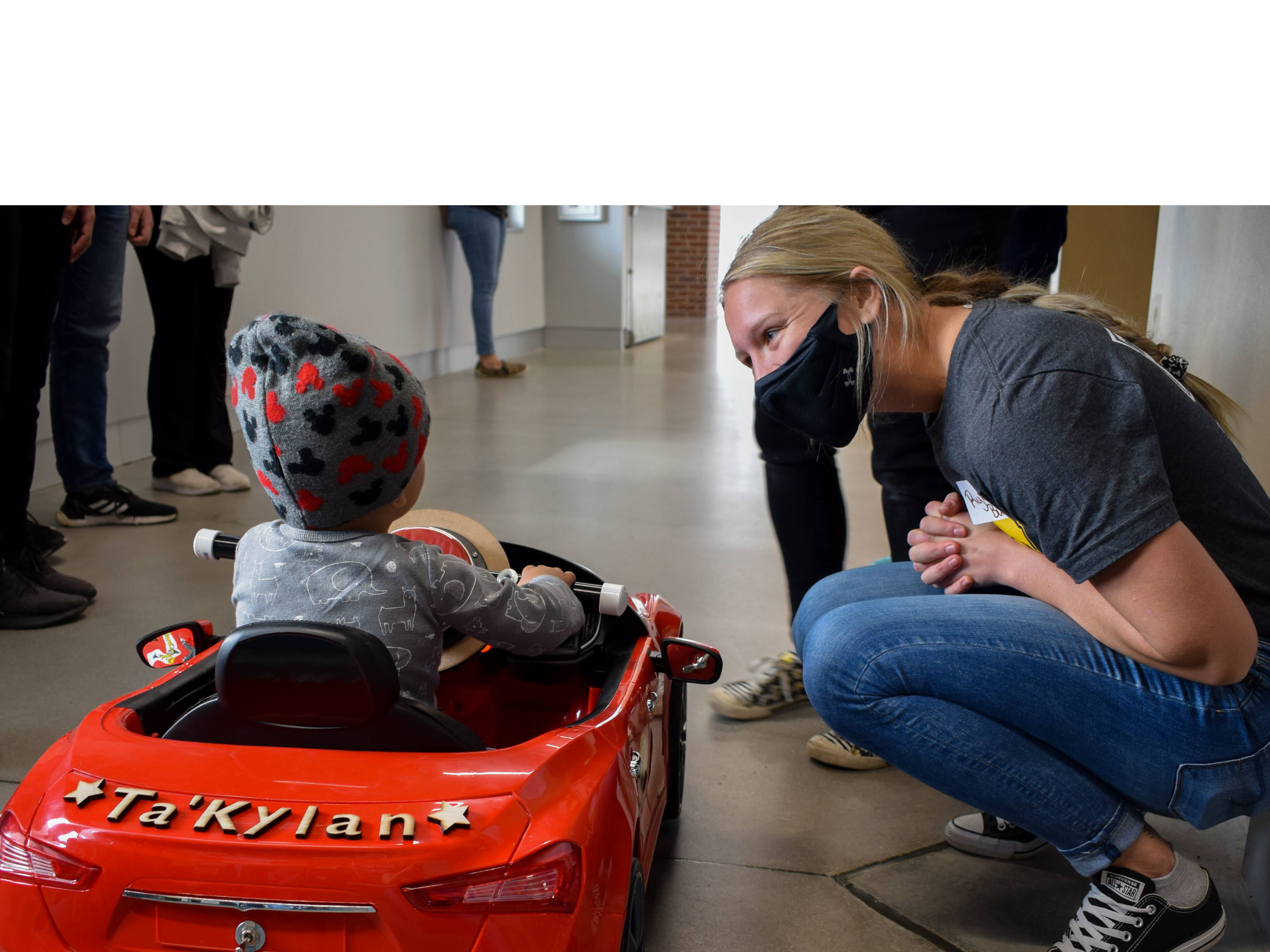 Students and staff form a pit crew to test the fit of a little girl to her new ride-on toy car from GoBabyGo.