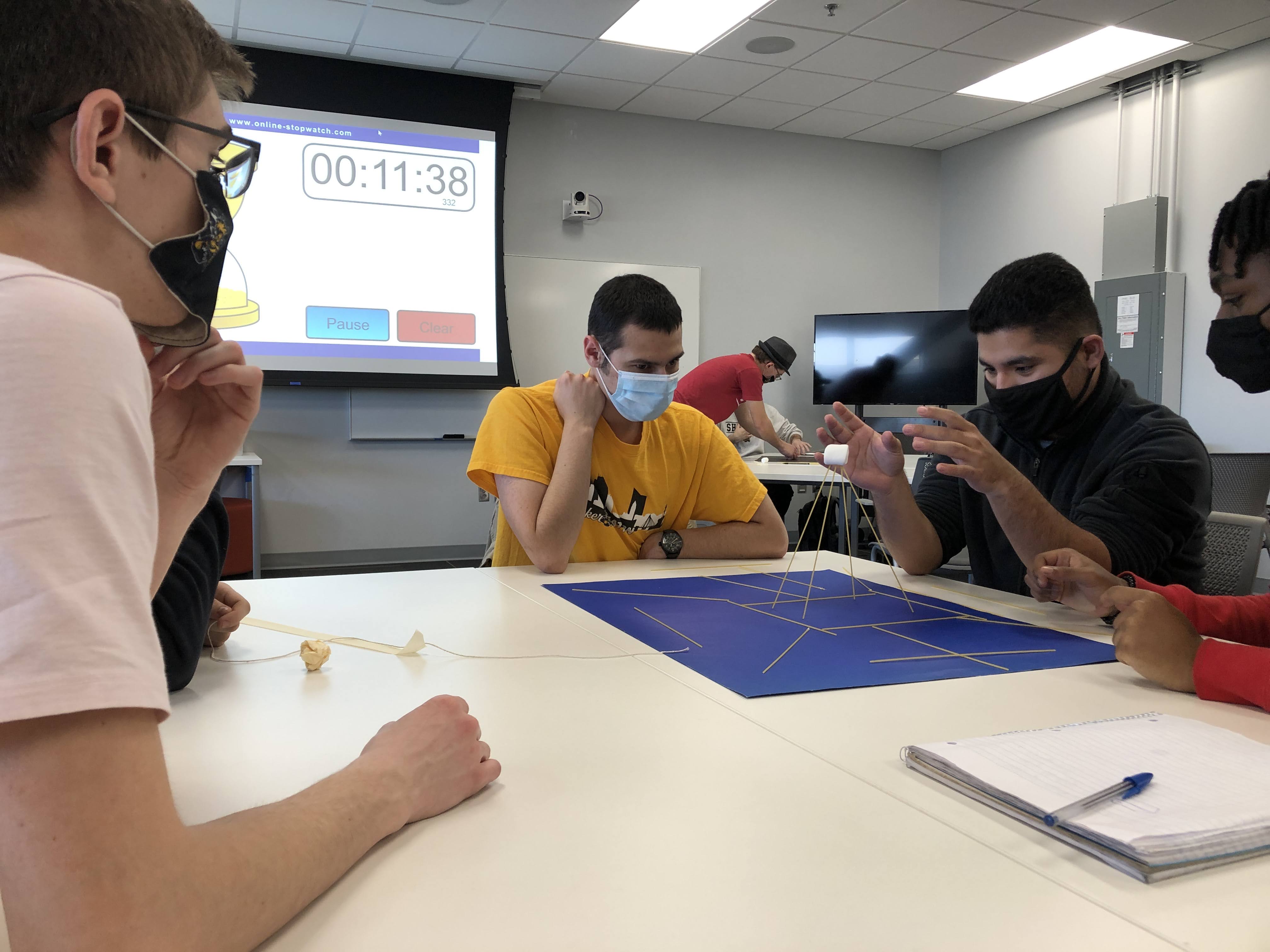Students working together to build a spaghetti tower with a timer counting down on the projector screen behind them.