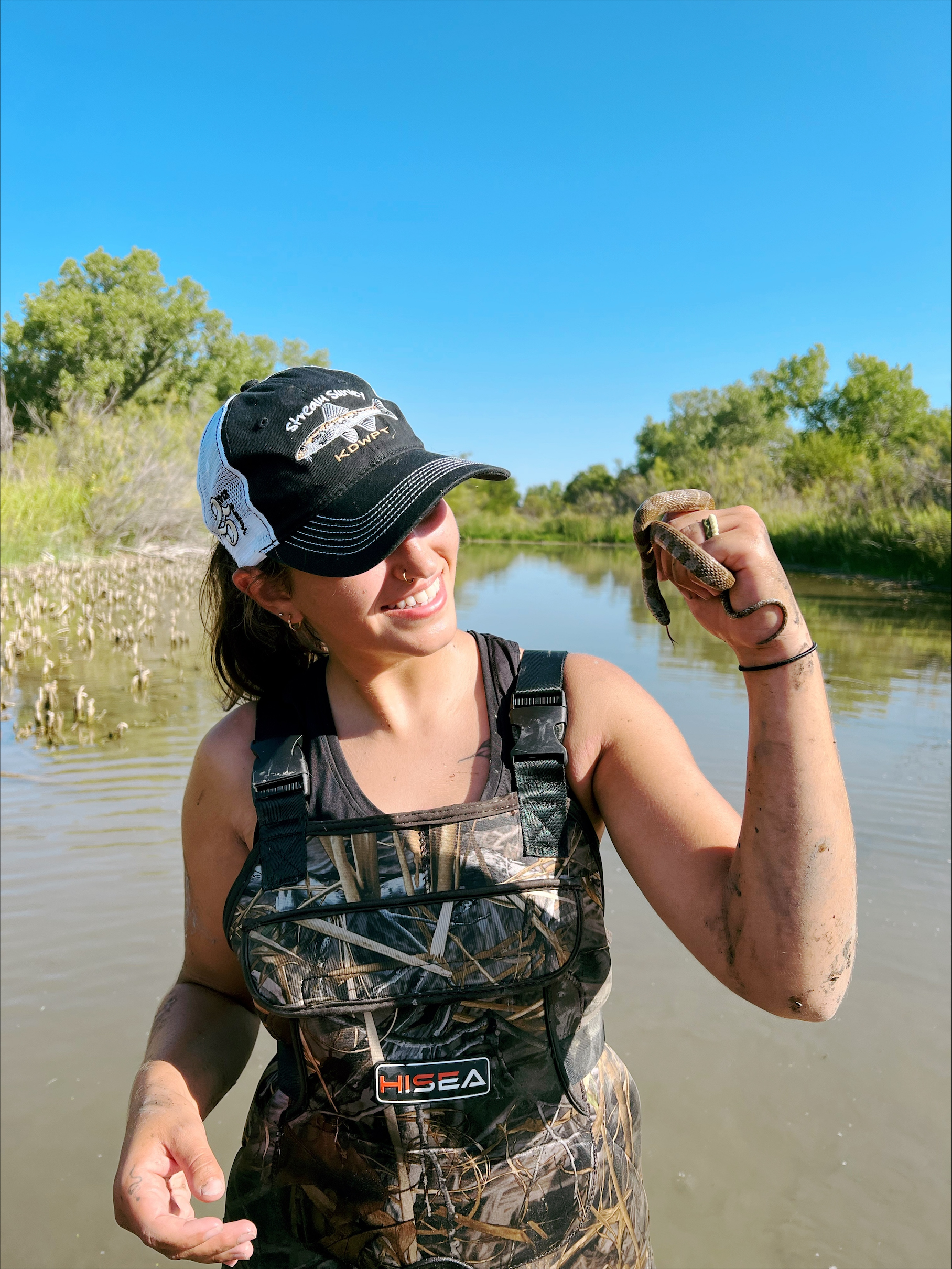 Student is near the camera wearing a baseball cap and waders. She is smiling but away from the camera, facing a small snake she is holding in her left hand.