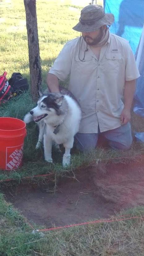 man kneeling by archaeological excavation wearing a fishing hat near a husky dog