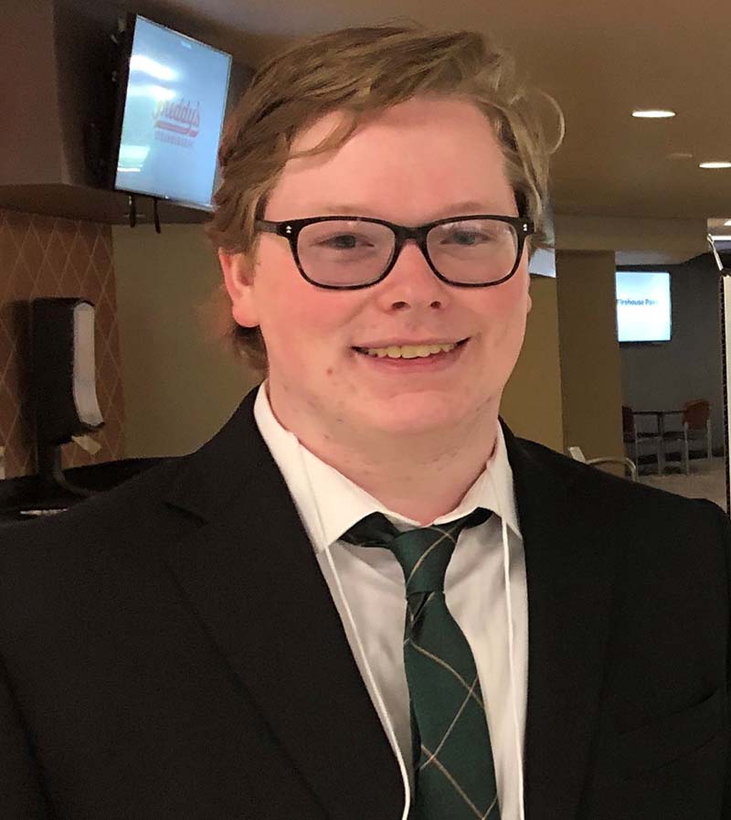 white man with blonde hair, smiling towards camera in black suit jacket and tie