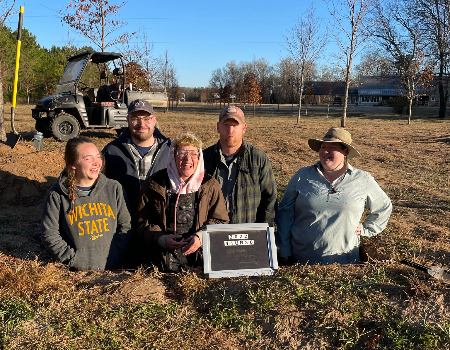 5 people standing in a square hole laughing. sign in front of them reads "41UR30 2022" 