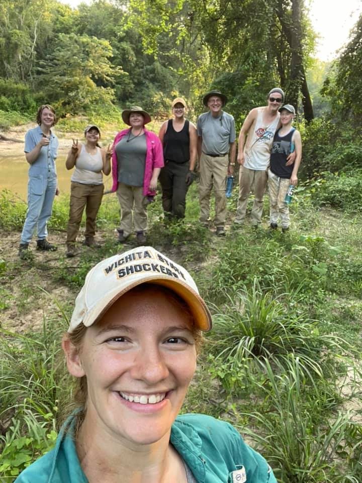 seven people standing in front of a river, dirty but smiling. A woman wearing a "Wichita State" hat smiles in the foreground.