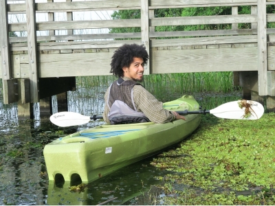 Student rowing in a kayak while surrounded by plants floating in the water.