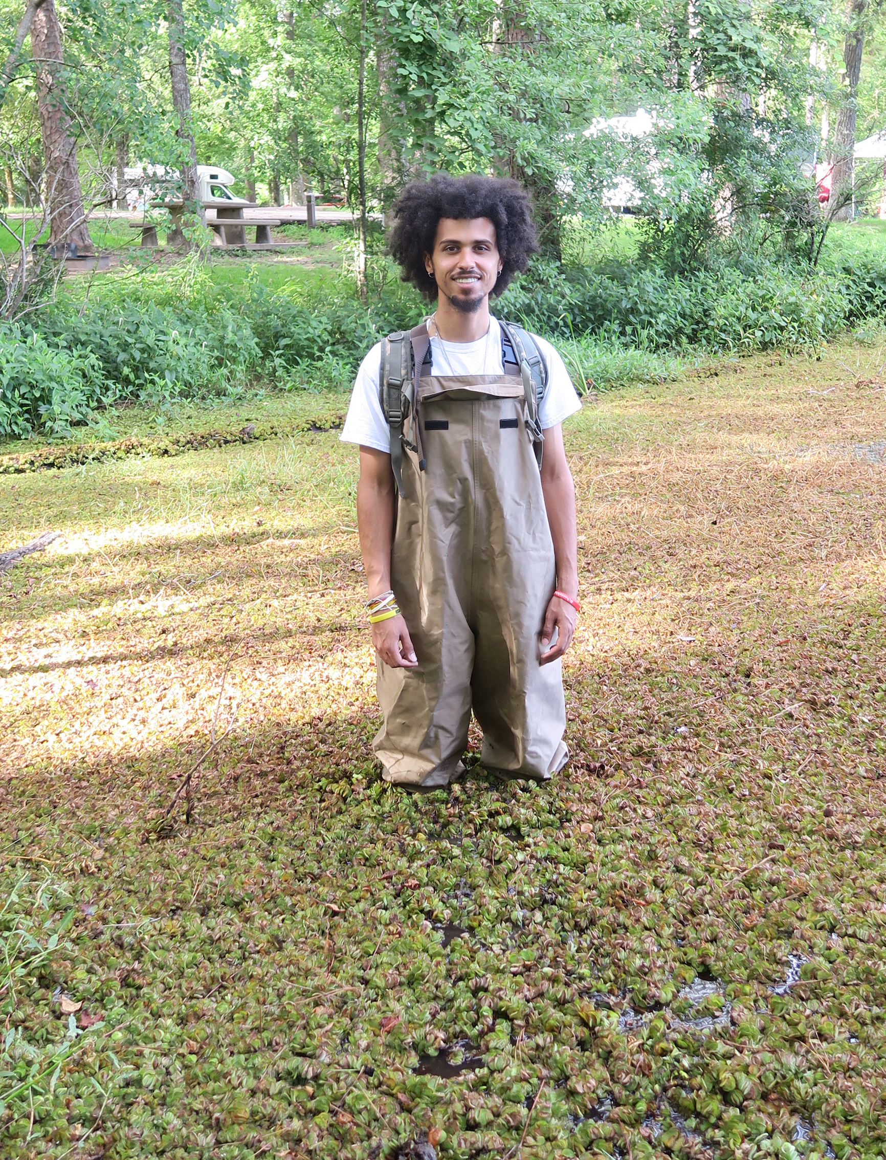 Photograph of student standing in water bog, wearing waders and surrounded by floating plant, Salvania. Student is smiling at the camera.