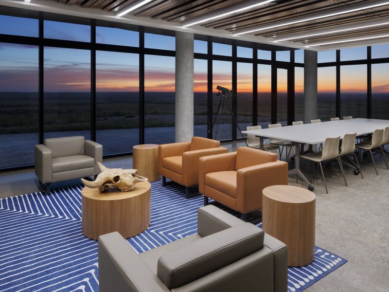 The interior lounge of the Youngmeyer Ranch building, cushioned seats surround a round table with a bull skull on it and a small flat table is lined with chairs on both sides. The walls are all windows, showing a view of the sunset
