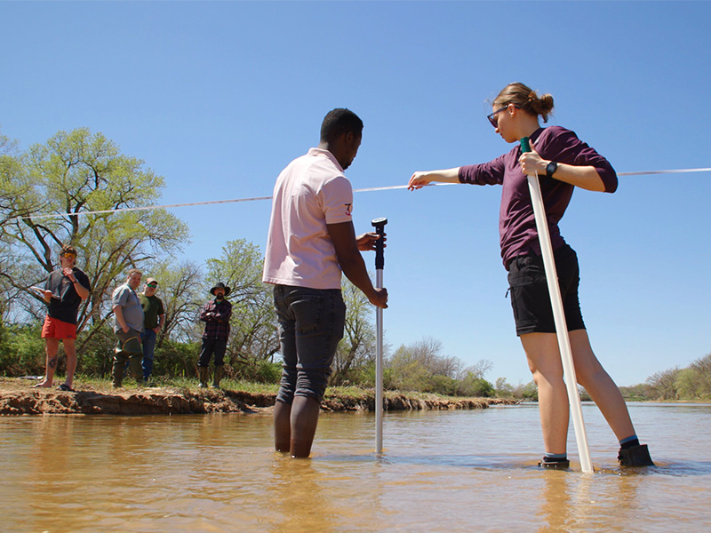 Students conduct measurements on a river. Two students are standing in the river while others take notes from the side.