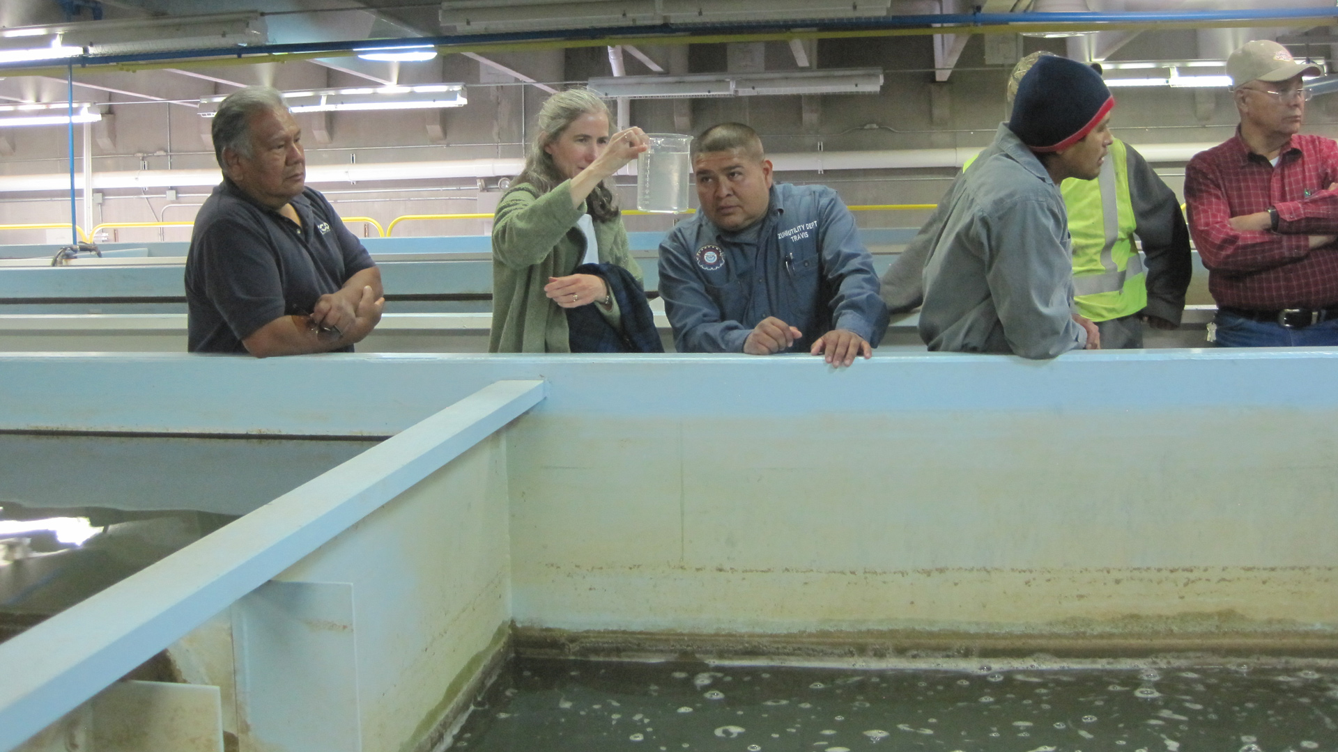 A woman holds up and examines a glass measuring cup full of water from a water treatment basin with two other staff from a Tribal water utility.