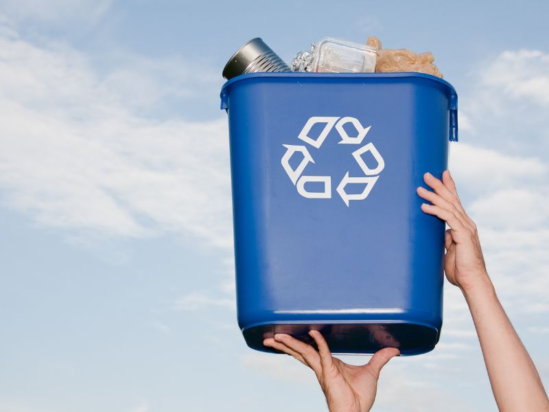 hands lifting up blue recycle bin filled with materials with blue sky background
