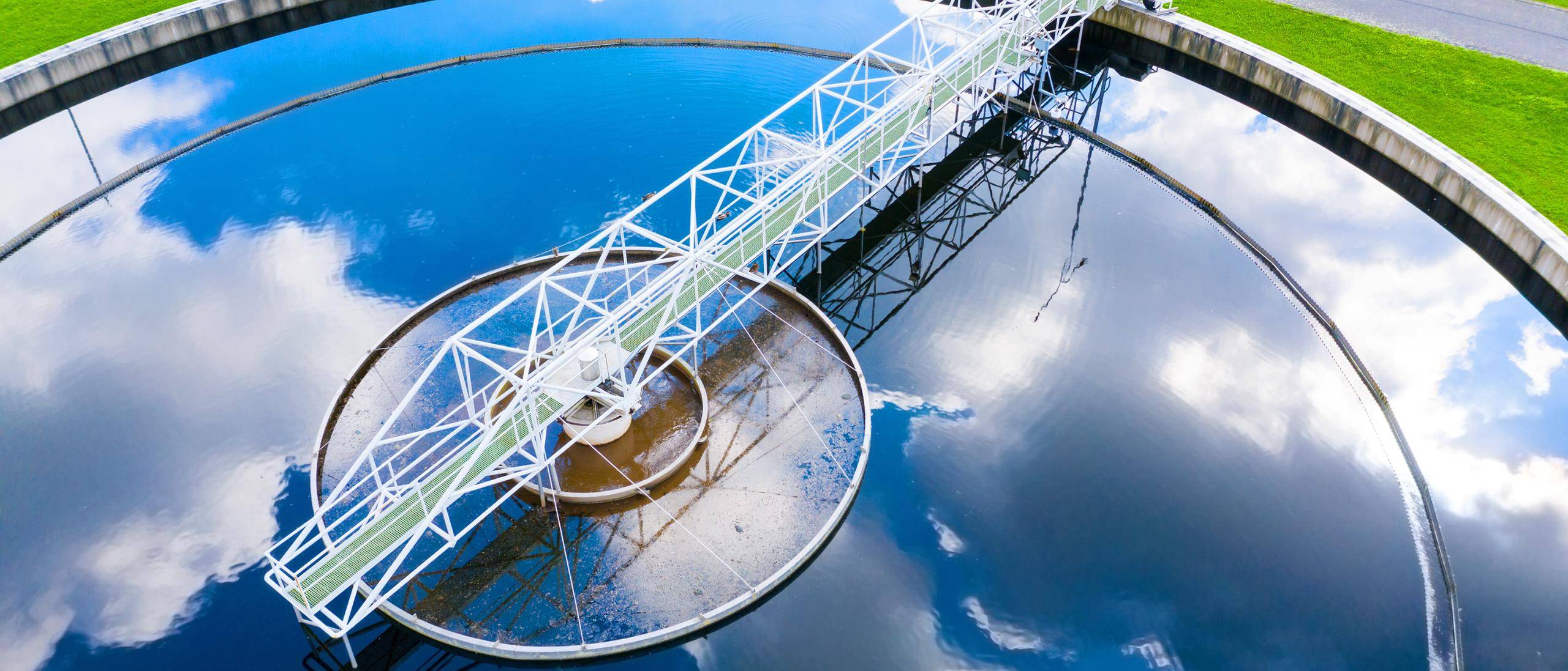 Overhead view of a water clarifier reflecting a blue sky with clouds sitting against vivid green grass.