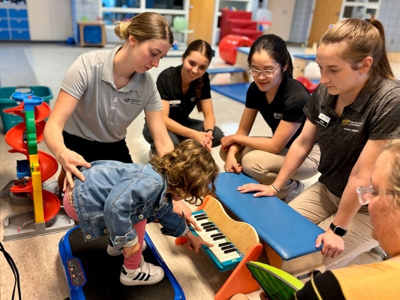 A child plays a toy piano while therapists observe and assist, with a vibration platform and toy ramp nearby.