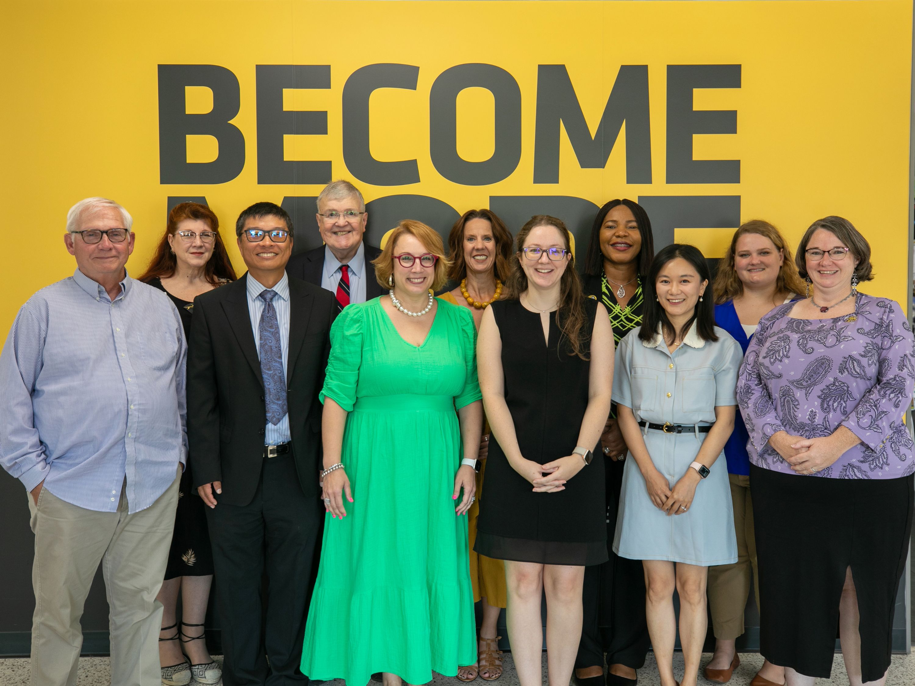 Hugo Wall School Faculty and Staff members stand in front of a large yellow wall with the word "BECOME" in large gray letters.