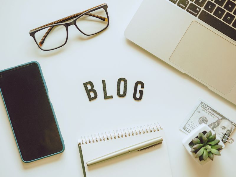 blog spelled out on table with glasses, laptop and hundred dollar bill paper clipped under a small deorative succulent plant